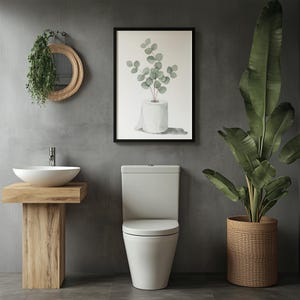 Op de afbeelding: Een moderne badkamer met een witte toilet, een houten wastafel met een witte wastafel en een ingelijste botanische print. Een grote plant in pot en een hangspiegel dragen bij aan de inrichting. De muren zijn neutraal grijs.