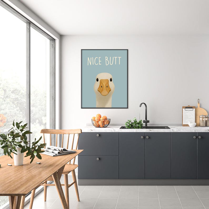 May include: A framed art print featuring a goose with a light blue background. The text "NICE BUTT" is above the goose. The goose has a yellow beak and black eyes. The print is displayed in a modern kitchen setting.