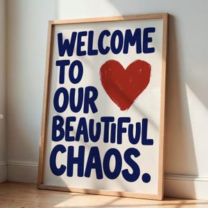 Könnte beinhalten: Ein gerahmter Kunstdruck mit dem dunkelblauen Text "WELCOME TO OUR BEAUTIFUL CHAOS." und einem großen, handgemalten roten Herz. Das Bild befindet sich in einem hellen Holzrahmen mit einem schlichten, klaren Design und einem einfachen Rand.