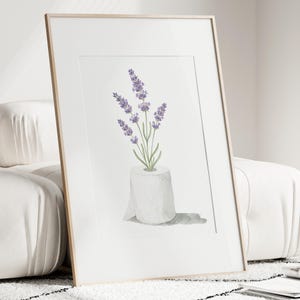 May include: A framed art print featuring a watercolor illustration of lavender sprigs in a roll of toilet paper. The lavender has purple flowers and green stems. The print is in a white mat and a light wood frame.