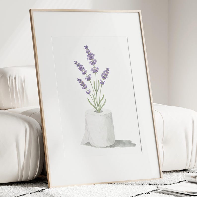 May include: A framed art print featuring a watercolor illustration of lavender sprigs in a roll of toilet paper. The lavender has purple flowers and green stems. The print is in a white mat and a light wood frame.