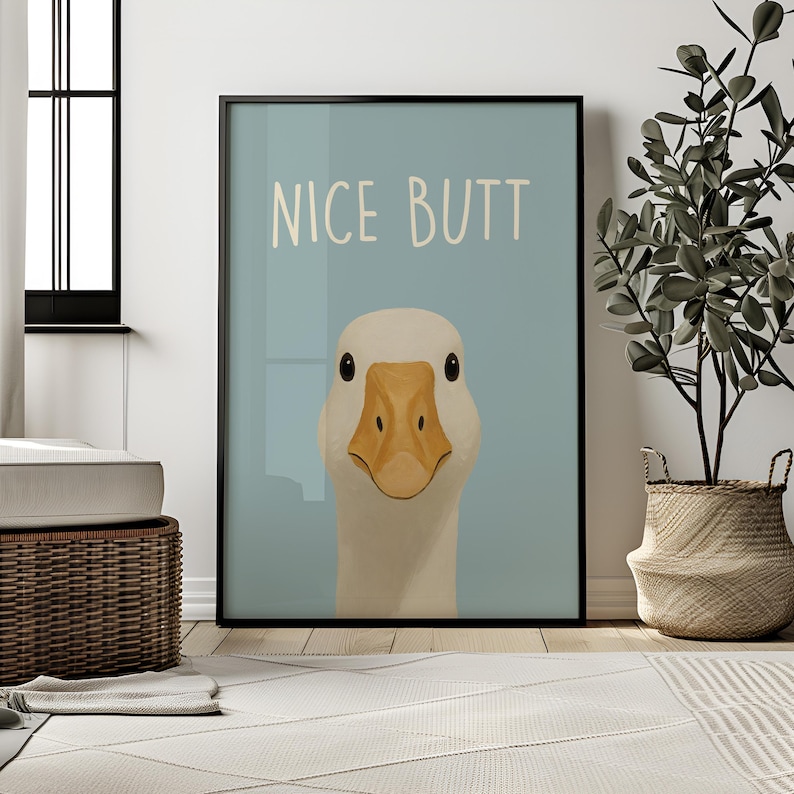 May include: Framed art print featuring a close-up illustration of a white goose with a yellow beak and the text "NICE BUTT" on a light blue background. The artwork is displayed in a black frame.