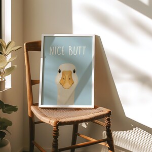 May include: A framed art print featuring a goose with the text "NICE BUTT" on a light blue background. The goose has a white head and neck with a yellow beak. The print is displayed on a wooden chair.