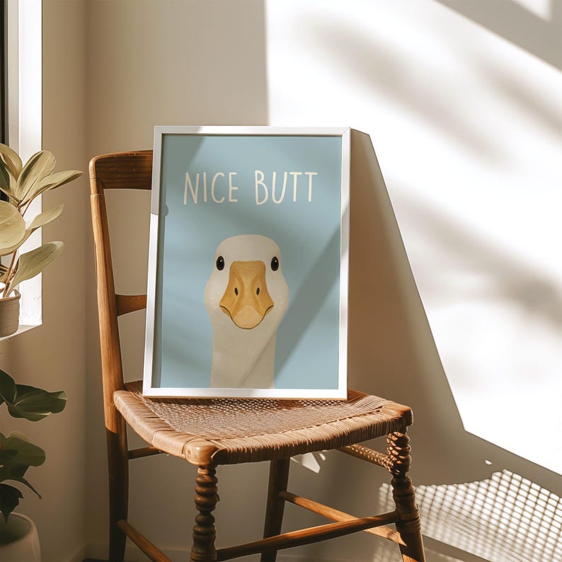 May include: A framed art print featuring a goose with the text "NICE BUTT" on a light blue background. The goose has a white head and neck with a yellow beak. The print is displayed on a wooden chair.