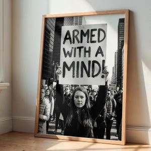 Könnte beinhalten: Schwarz-Weiß-Druck in einem Rahmen, der eine Menschenmenge und eine Frau mit einem Schild mit der Aufschrift "ARMED WITH A MIND!" zeigt. Der Druck befindet sich in einem Holzrahmen und lehnt an einer Wand.
