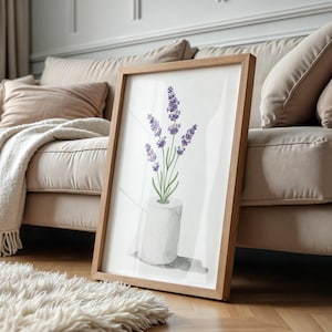 May include: A framed watercolor print featuring lavender flowers in a white vase. The artwork has a light brown wooden frame and is leaning against a beige sofa. The image is set on a hardwood floor with a fluffy white rug.