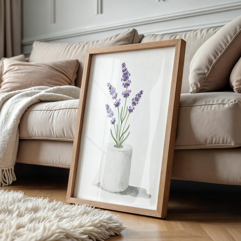 May include: A framed watercolor print featuring lavender flowers in a white vase. The artwork has a light brown wooden frame and is leaning against a beige sofa. The image is set on a hardwood floor with a fluffy white rug.