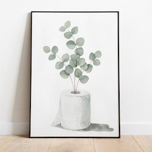 Op de afbeelding: Waterverfprint van een rol wit toiletpapier met een takje eucalyptus. De eucalyptus heeft groene bladeren en rode stelen. Het kunstwerk is ingelijst in een zwart frame.