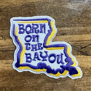 May include: Embroidered patch in the shape of Louisiana, with the words "BORN ON THE BAYOU" in purple lettering. The patch has a white background, a yellow outline, and a purple border.