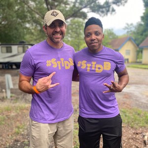 May include: Two people wearing purple t-shirts with the text "#STDB" in yellow. The person on the left is wearing a tan baseball cap.