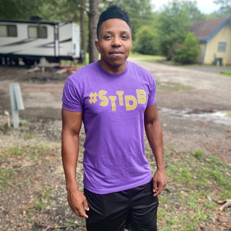 May include: A person wearing a purple t-shirt with the text "#STDB" in yellow. The person is standing in front of a green grassy area.