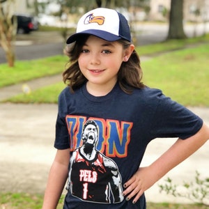 May include: A young person wearing a navy blue t-shirt with a basketball player graphic and the text "Zion" on the front. The shirt also features the text "Pels 1". The person is also wearing a white and blue baseball cap with a bird logo.