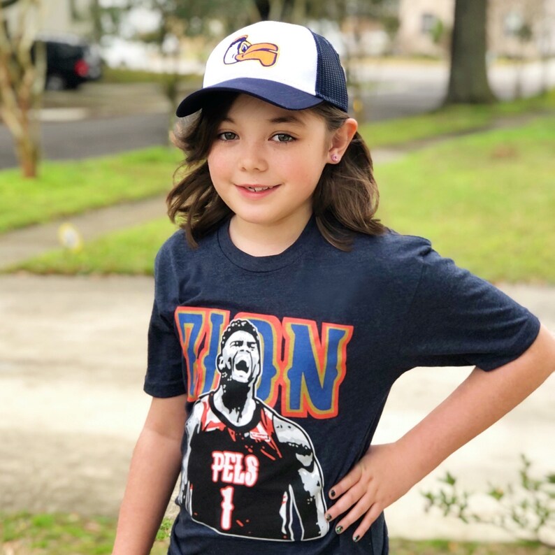 May include: A young person wearing a navy blue t-shirt with a basketball player graphic and the text "Zion" on the front. The shirt also features the text "Pels 1". The person is also wearing a white and blue baseball cap with a bird logo.