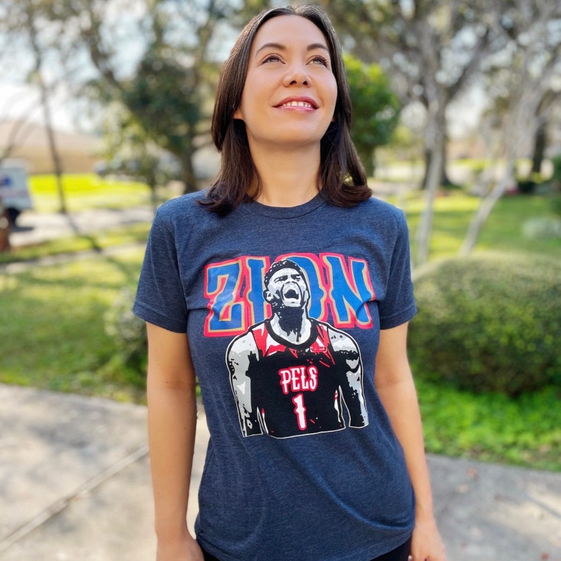 May include: A navy blue t-shirt with a graphic of basketball player Zion Williamson wearing a red and white jersey with the text "PELS 1" on it. The text "ZION" is printed in blue and red letters above the graphic.