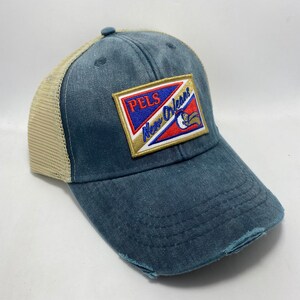 May include: A blue and tan distressed trucker hat with a patch that says "PELS New Orleans" in gold lettering on a blue, red, and white background.