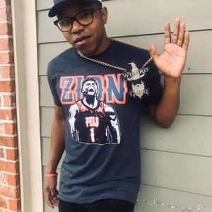 May include: A man wearing a navy blue t-shirt with a white and red graphic of a basketball player with the text "Zion" and "Pels 1". He is also wearing a black and white baseball cap with a red logo and a gold chain with a pendant that says "Wizard Gang".