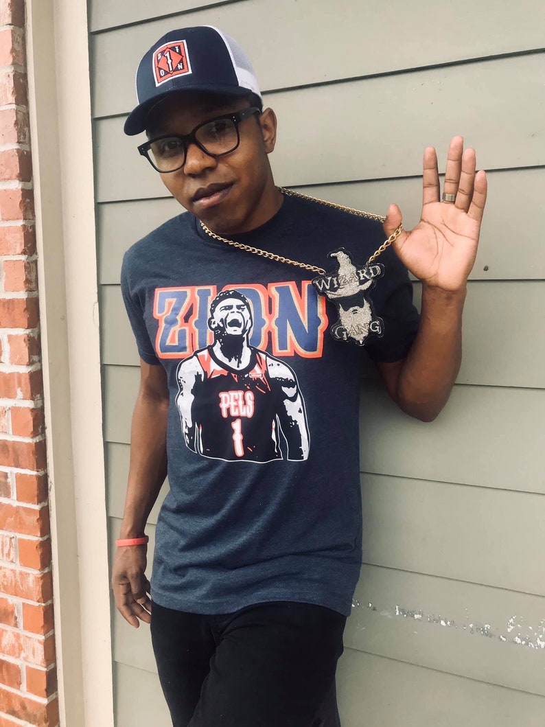May include: A man wearing a navy blue t-shirt with a white and red graphic of a basketball player with the text "Zion" and "Pels 1". He is also wearing a black and white baseball cap with a red logo and a gold chain with a pendant that says "Wizard Gang".