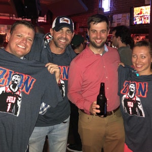 May include: Four people are wearing blue t-shirts with a basketball player graphic and the text "Zion" on the front. One person is holding a bottle of beer.