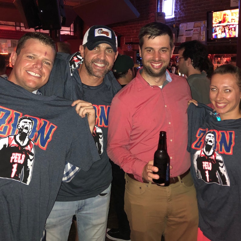 May include: Four people are wearing blue t-shirts with a basketball player graphic and the text "Zion" on the front. One person is holding a bottle of beer.