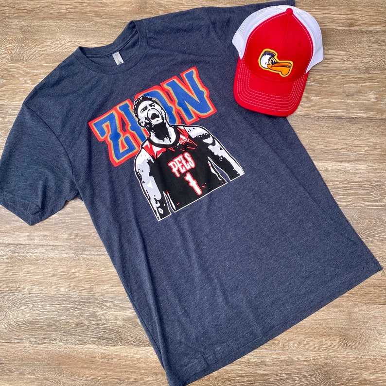 May include: A navy blue t-shirt with a graphic of a basketball player in a red, white, and blue jersey with the word "ZION" in blue and white letters. The player is wearing a red headband and has a determined expression on his face. The jersey has the word "PELS" and the number "1" on it. A red and white baseball cap with a duck logo is also in the image.