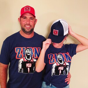 May include: Two people wearing navy blue t-shirts with a graphic of Zion Williamson, a basketball player for the New Orleans Pelicans. The shirts have the text "Zion" in large letters. The people are wearing red and blue baseball caps with the letter "H" on them.