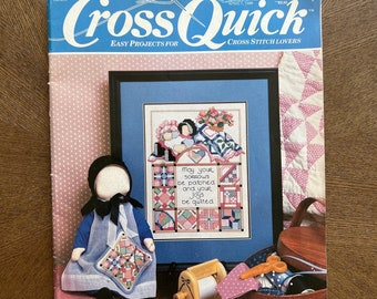 May include: A vintage issue of "Cross Quick" magazine with a blue and white cover. The magazine features cross-stitch projects, with a framed cross-stitch design visible. A doll in a blue dress and bonnet is in the foreground, along with sewing supplies.