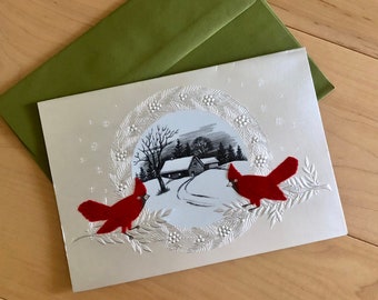May include: A holiday greeting card with a winter scene and two red cardinals perched on a branch. The card has a silver finish and is accompanied by a green envelope. The winter scene depicts a snowy landscape with houses and trees.