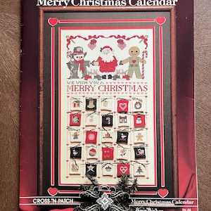 May include: A cross-stitch pattern book titled "Merry Christmas Calendar." The cover features a framed cross-stitch design with a snowman, Santa Claus, and a gingerbread man. The calendar has 24 small squares with Christmas-themed images.
