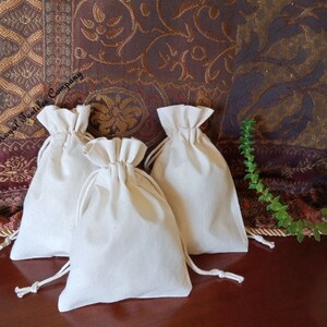 May include: Three white cotton drawstring bags on a brown surface. The bags are made of natural fabric and have a simple design.