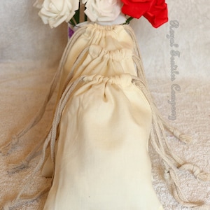 May include: Three natural cotton drawstring bags with a white background and beige drawstrings. The bags are stacked on top of each other and are sitting on a white fluffy surface. The text "Royal Textiles Company" is visible in the upper right corner of the image.