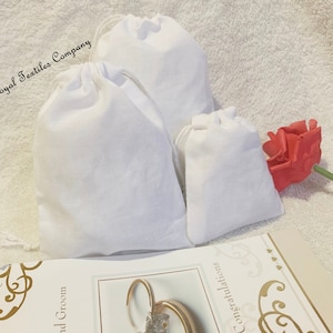 May include: Three white cotton drawstring bags of varying sizes. The largest bag has the text "Royal Textiles Company" printed on it. The bags are on a white fluffy surface.