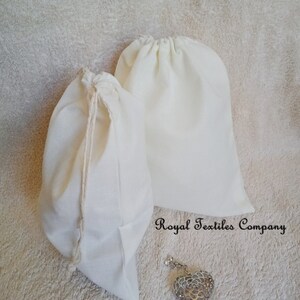 May include: Two cream-coloured drawstring bags made of a soft fabric. The bags are different sizes, with one slightly smaller than the other. A silver heart-shaped pendant with intricate detailing rests near the bags. The text "Royal Textiles Company" is visible.