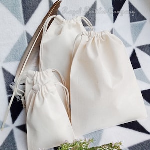 May include: A set of four white cotton drawstring bags on a grey and white patterned surface. The bags are made of natural cotton fabric and have a simple design. The bags are perfect for packaging small gifts or favours.