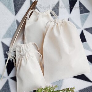 May include: Four white cotton drawstring bags of various sizes. The bags are on a grey and white patterned fabric background. The text "Royal Textiles Company" is visible in the top right corner of the image.