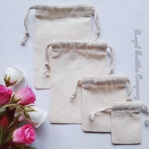 May include: Four white cotton drawstring pouches of varying sizes. The pouches are arranged in a row from largest to smallest. The pouches are made of natural cotton fabric and have a simple, elegant design.