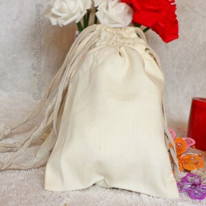 May include: A set of 10 natural cotton muslin drawstring bags with a white background and natural cotton drawstrings. The bags are shown on a white fluffy surface with red, white, and pink flowers in the background.