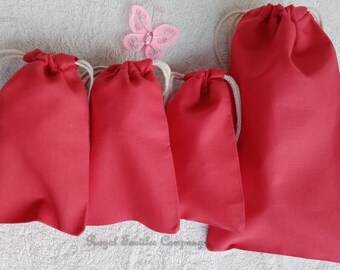 May include: Five red drawstring bags made of fabric with white drawstrings. The bags are arranged in a row on a white fuzzy surface.