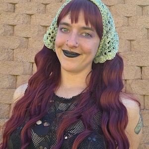 May include: A light green crocheted head covering with a scalloped edge. The head covering is worn over long, wavy, auburn hair. The person is wearing a black mesh top.