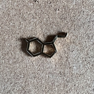 May include: A gold-colored serotonin molecule charm, a chemical messenger in the brain, is displayed on a textured, light brown corkboard surface. The charm's geometric design is clearly visible, showcasing its unique shape and structure.