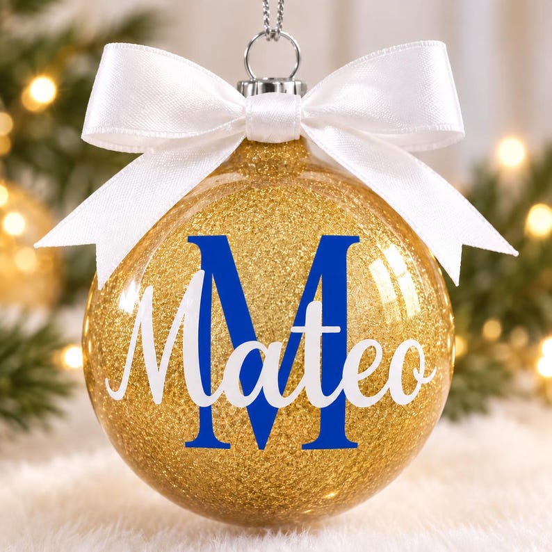 May include: A clear spherical Christmas ornament filled with gold glitter, featuring a large blue letter M and the name Mateo in white script. The ornament is topped with a silver cap and a tied white satin ribbon bow for hanging.