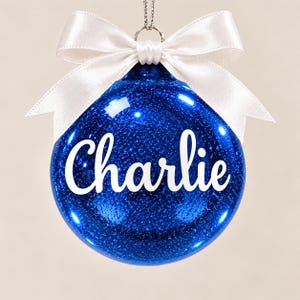 May include: A round, blue glittery Christmas ornament featuring the name Charlie written in white script lettering across the center. The ornament is topped with a white satin ribbon tied into a bow and includes a silver hanging loop at the top.