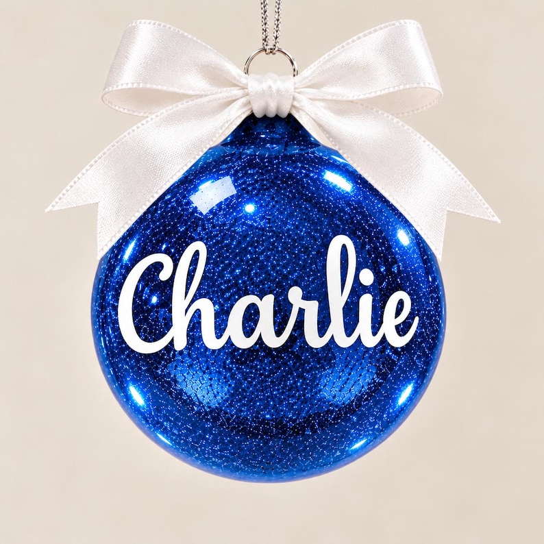 May include: A round, blue glittery Christmas ornament featuring the name Charlie written in white script lettering across the center. The ornament is topped with a white satin ribbon tied into a bow and includes a silver hanging loop at the top.