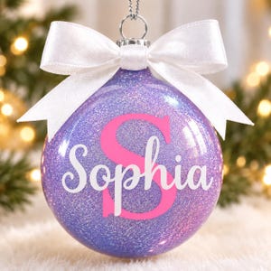 May include: A spherical purple glitter Christmas ornament featuring a large pink letter S and the name Sophia written in white cursive script. The ornament is topped with a silver metal cap and a tied white satin ribbon bow for hanging.