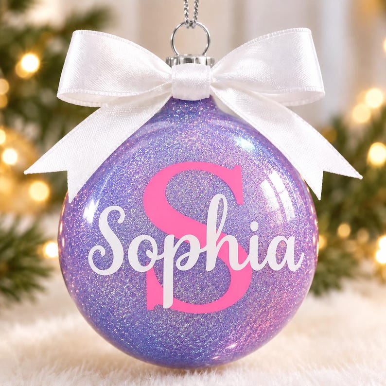 May include: A spherical purple glitter Christmas ornament featuring a large pink letter S and the name Sophia written in white cursive script. The ornament is topped with a silver metal cap and a tied white satin ribbon bow for hanging.