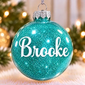 May include: A round, clear glass Christmas ornament filled with vibrant teal glitter. The name Brooke is printed in white, cursive lettering across the center. A silver metal cap with a snowflake pattern sits at the top with a hanging loop.