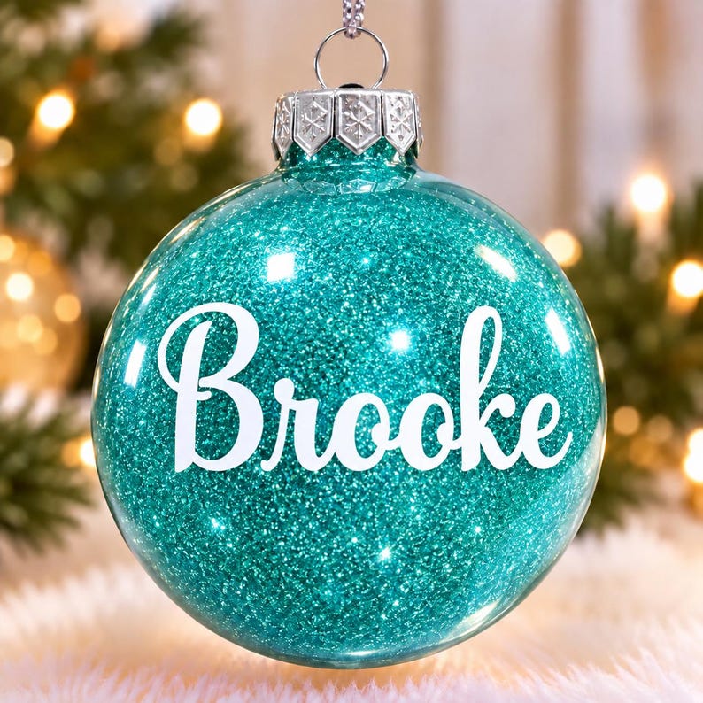 May include: A round, clear glass Christmas ornament filled with vibrant teal glitter. The name Brooke is printed in white, cursive lettering across the center. A silver metal cap with a snowflake pattern sits at the top with a hanging loop.