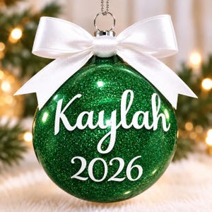 May include: A spherical green glitter Christmas ornament featuring the white text "Kaylah" in a script font and "2026" in a serif font. The ornament is topped with a silver metal cap and a white satin ribbon tied into a decorative bow.
