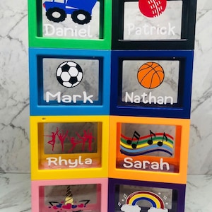 May include: Six colourful, clear plastic boxes with different images and names on each. The boxes are stacked in two rows of three. The top row has a blue truck with the name "Daniel", a red cricket ball with the name "Patrick", a football with the name "Mark", and a basketball with the name "Nathan". The bottom row has a group of dancers with the name "Rhyla", a rainbow with musical notes with the name "Sarah", a unicorn with the name "Brooke", and a rainbow with clouds with the name "Katie".