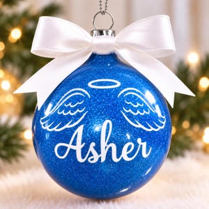 May include: A round blue glitter Christmas ornament featuring a white vinyl decal of angel wings and a halo above the name Asher in script font. The ornament is topped with a silver metal cap and a tied white satin ribbon bow for hanging.