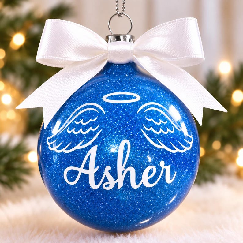 May include: A round blue glitter Christmas ornament featuring a white vinyl decal of angel wings and a halo above the name Asher in script font. The ornament is topped with a silver metal cap and a tied white satin ribbon bow for hanging.